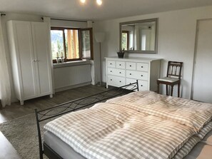 Une chambre à coucher avec un lit, une commode, une chaise, un radiateur et une fenêtre avec des rideaux.