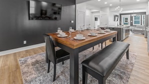 Un comedor moderno con una larga mesa de madera dispuesta para seis comensales, sillas negras y un banco. Un televisor de pantalla plana está montado en la pared oscura sobre la mesa.