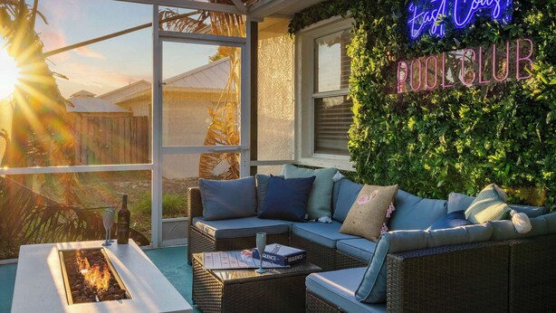Un espacio al aire libre iluminado por el sol con zona de estar, un letrero de neón que dice 'East Coast Pool Club', un elemento decorativo de fuego y un cerramiento de cristal.