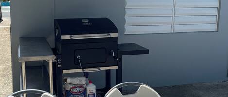 A barbecue grill with a table and chairs in front, a window with blinds, and a corrugated metal roof.