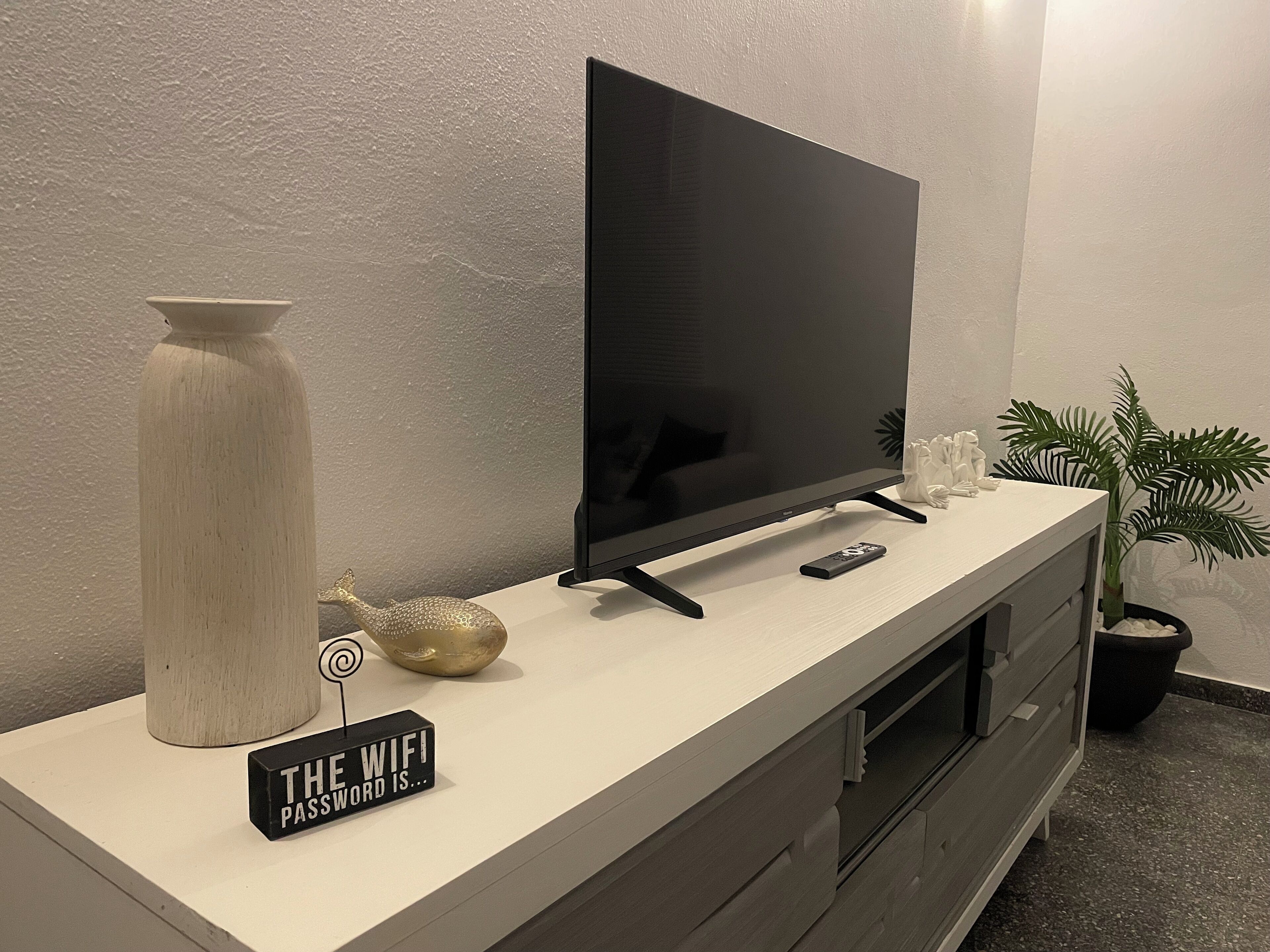 Una sala de estar moderna con un televisor de pantalla plana, un florero, una caracola decorativa, un control remoto y un cartel que dice 'THE WIFI PASSWORD IS...'.