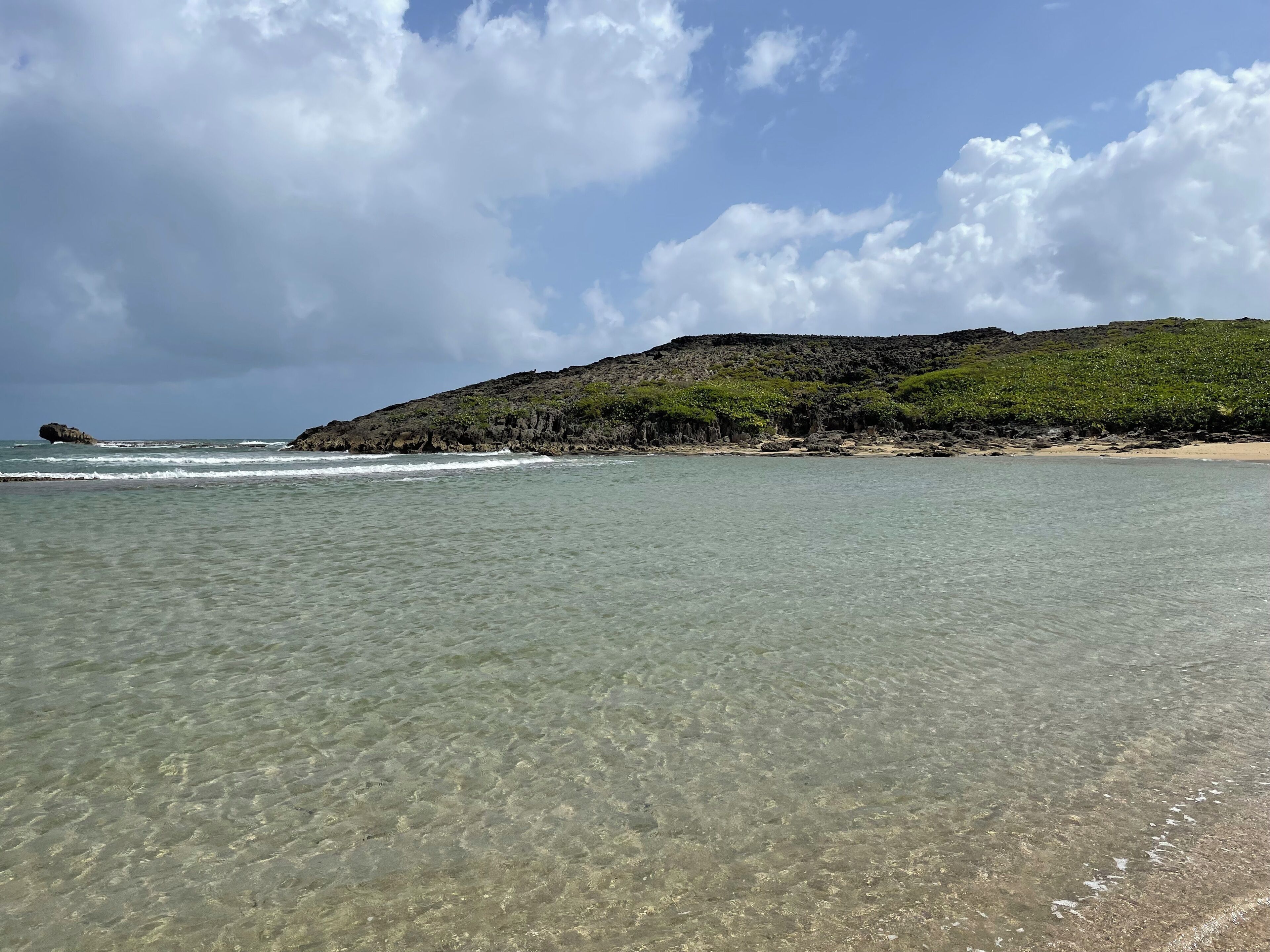 Una playa con aguas transparentes y poco profundas, un litoral rocoso y una colina con vegetación.