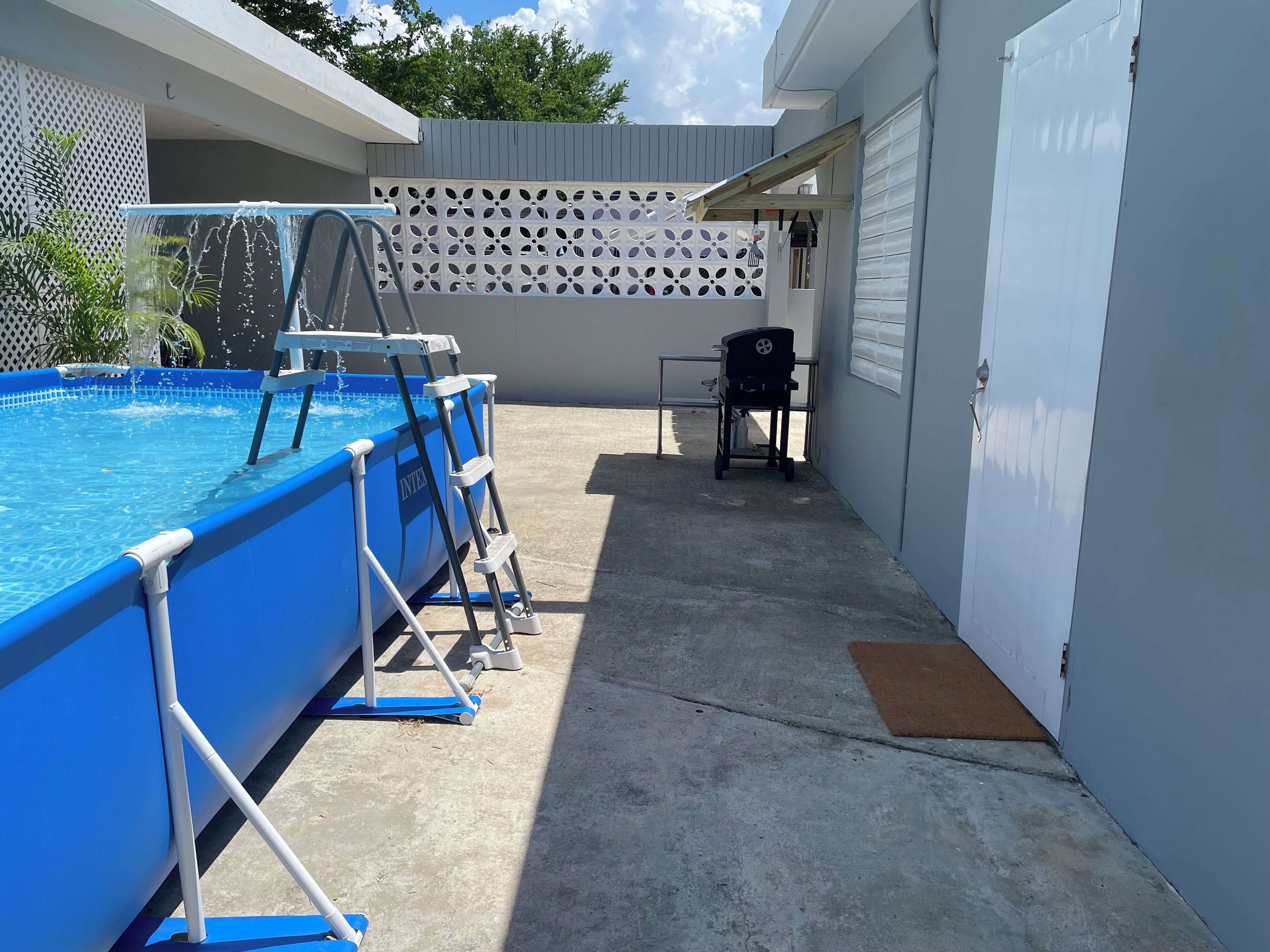 An outdoor swimming pool with a ladder, a barbecue grill, and a covered area with a concrete floor.