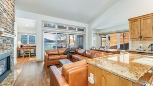 A spacious living area with a large leather sofa, a stone fireplace, and a kitchen with wooden cabinets and granite countertops. - New! 30-Day Min | Whitefish Lake Views | 3BR Condo w/ Deck Near Downtown (Whitefish)