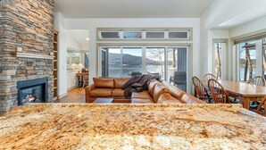 A spacious living room with a stone fireplace, a large sectional sofa, and a dining area with a view of the outdoors. - New! 30-Day Min | Whitefish Lake Views | 3BR Condo w/ Deck Near Downtown (Whitefish)