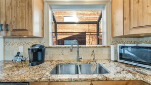 A kitchen with granite countertops, stainless steel sinks, wooden cabinets, a built-in microwave, and a window with a view of the outside. - New! 30-Day Min | Whitefish Lake Views | 3BR Condo w/ Deck Near Downtown (Whitefish)