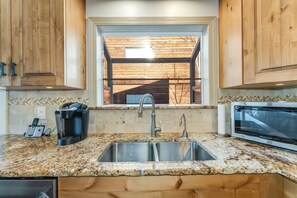 A kitchen with granite countertops, stainless steel sinks, wooden cabinets, a built-in microwave, and a window with a view of the outside. - New! 30-Day Min | Whitefish Lake Views | 3BR Condo w/ Deck Near Downtown (Whitefish)