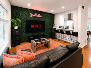 Una sala de estar moderna con un televisor de pantalla plana, una mesa de fútbol de mesa, un sofá y una zona de cocina al fondo.