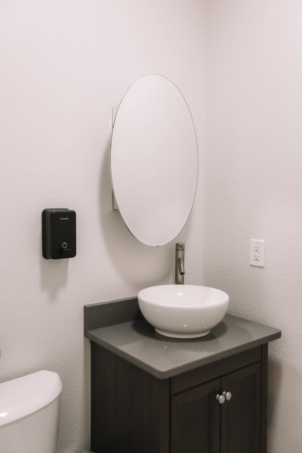 A bathroom with a white toilet, a dark vanity with a white sink, and a wall-mounted oval mirror. - 2 Homes, 8BR | Sleeps 30 | Game Room & Fire Pit (Denver)