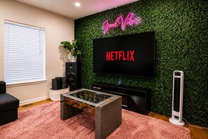 Una sala de estar con una televisión de pantalla plana montada en una pared verde, una mesa de fútbol de mesa, un sofá negro y una alfombra rosa.