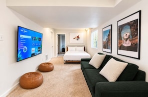 A modern bedroom with a large flat-screen TV, a bed, a green sofa, and two leather poufs.