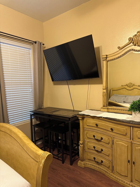 A room with a bed, a desk with two chairs, a television mounted on the wall, and a wooden dresser.