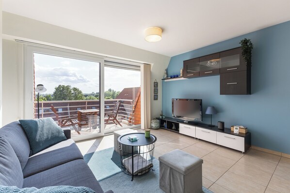 Un salon moderne comprenant un canapé, une petite table ronde, un téléviseur et une porte-fenêtre coulissante donnant sur un balcon aménagé avec du mobilier d’extérieur.
