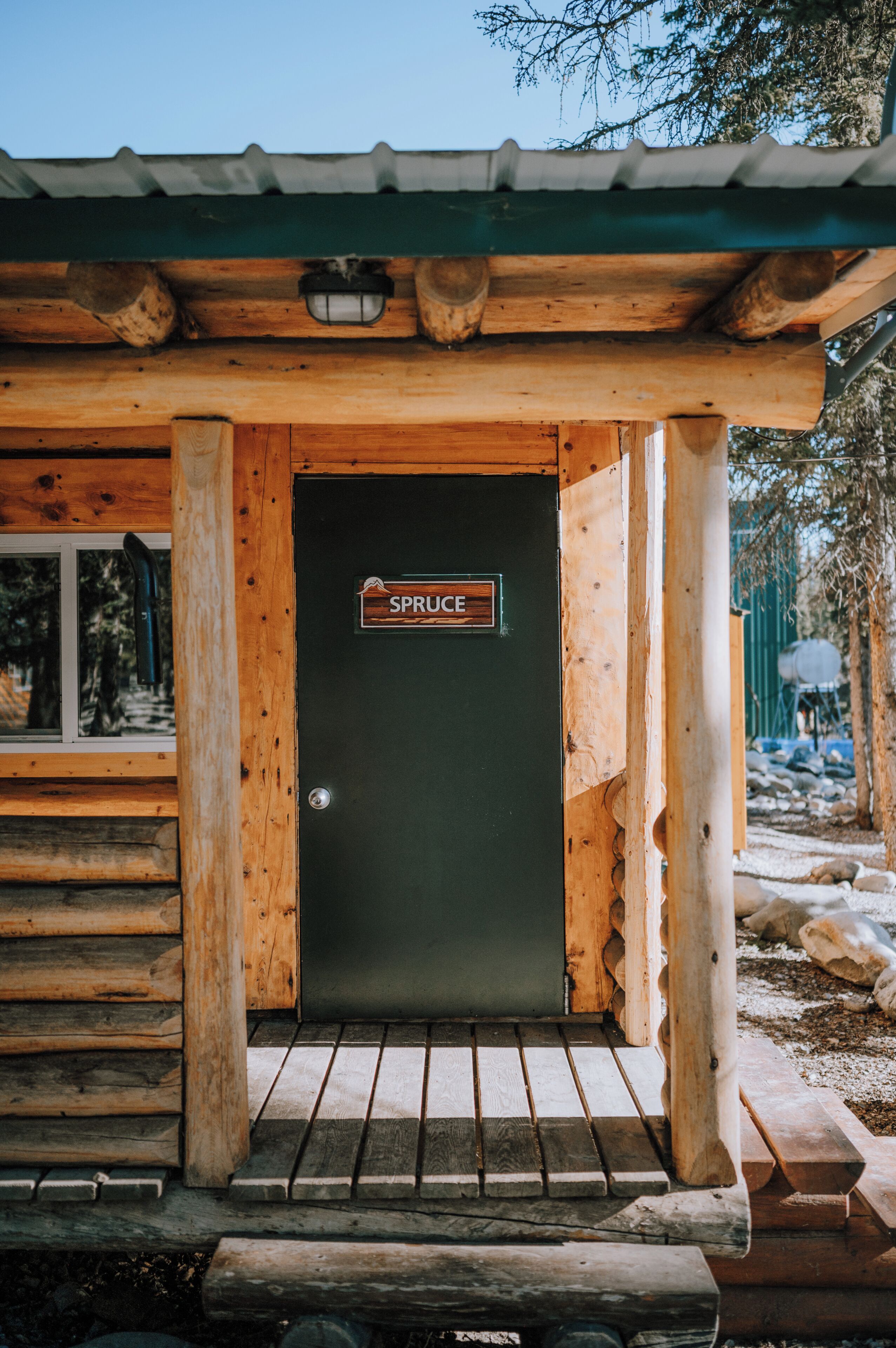 A wooden cabin with a sign reading 'SPRUCE' at the entrance, a window, and a metal roof.