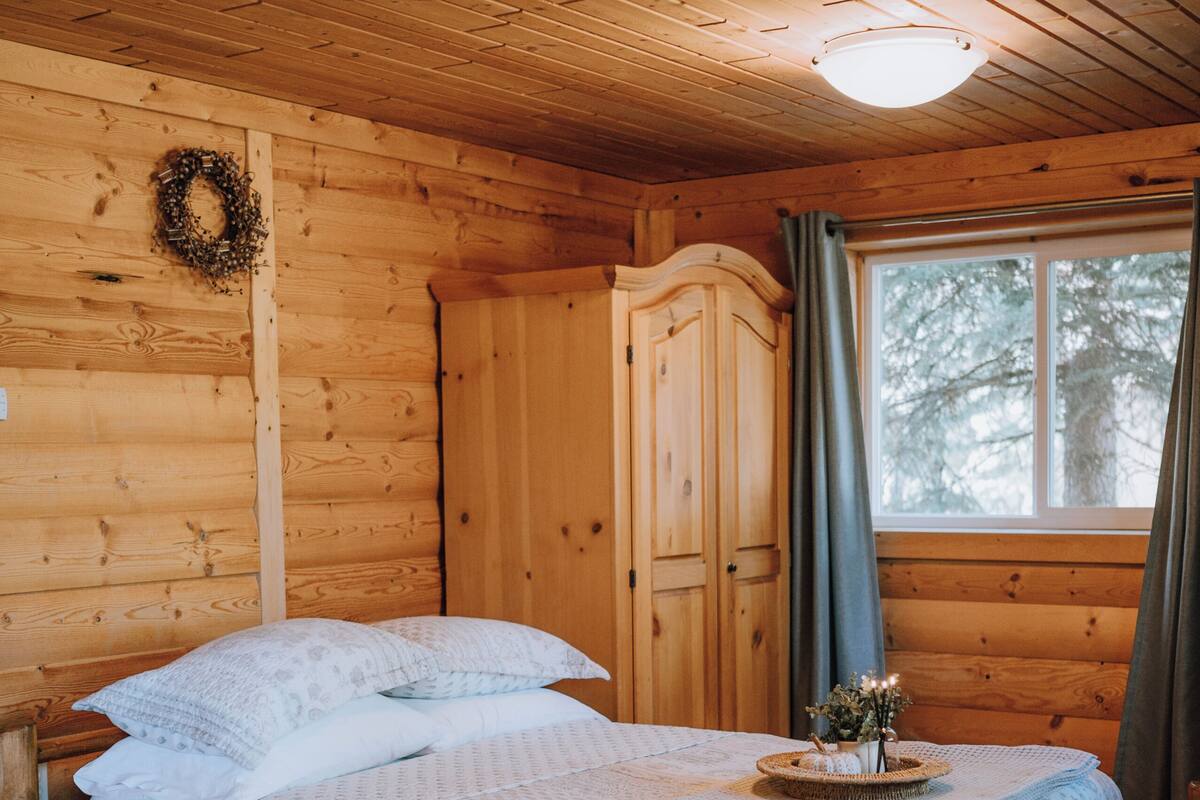 Ein Schlafzimmer einer Holzhütte mit einem Bett, einem Kleiderschrank, einem Fenster mit Vorhängen und einem Kranz an der Wand.
