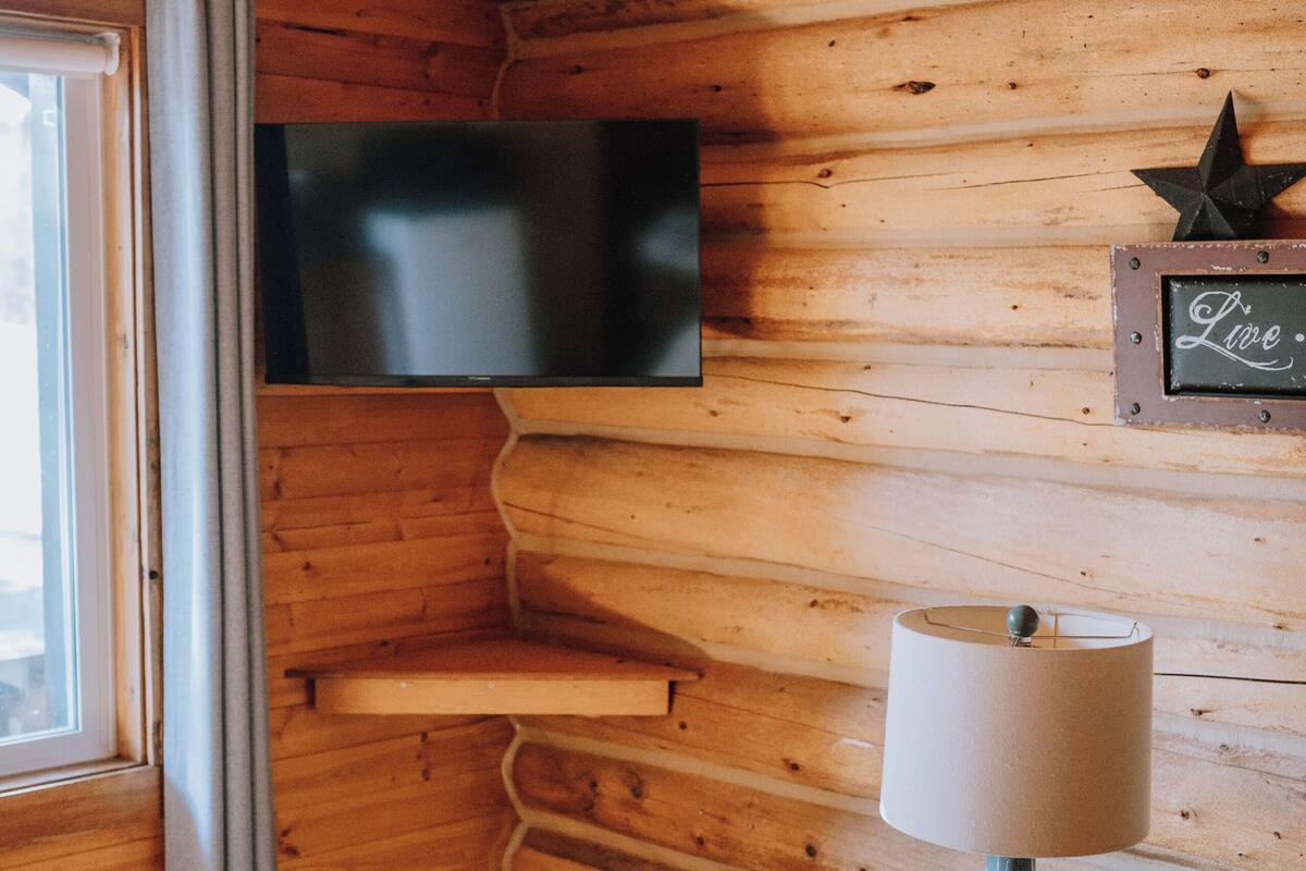 Ein gemütliches Blockhauszimmer mit Holzwänden, einem an der Wand befestigten Fernseher, einer Lampe, einer Kerze und einer Lederbank.