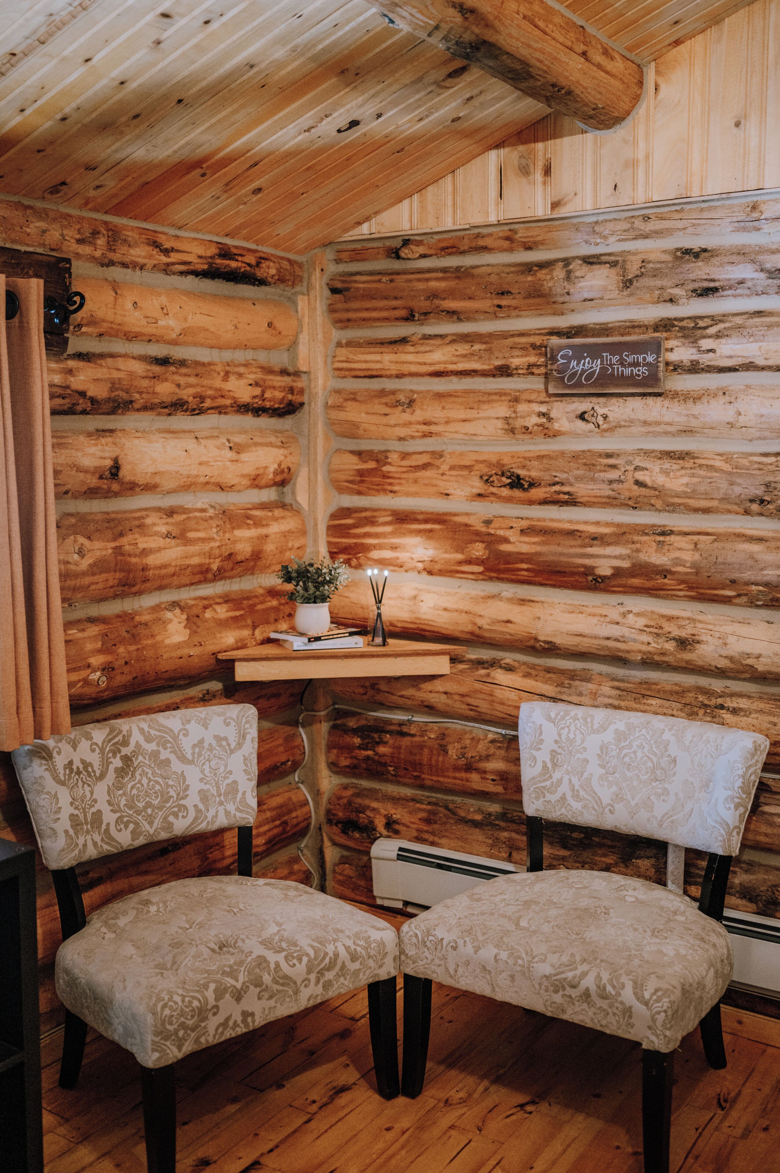 Interior de uma cabana de tronco com duas poltronas estofadas, uma pequena mesa com uma planta em vaso e um letreiro na parede.