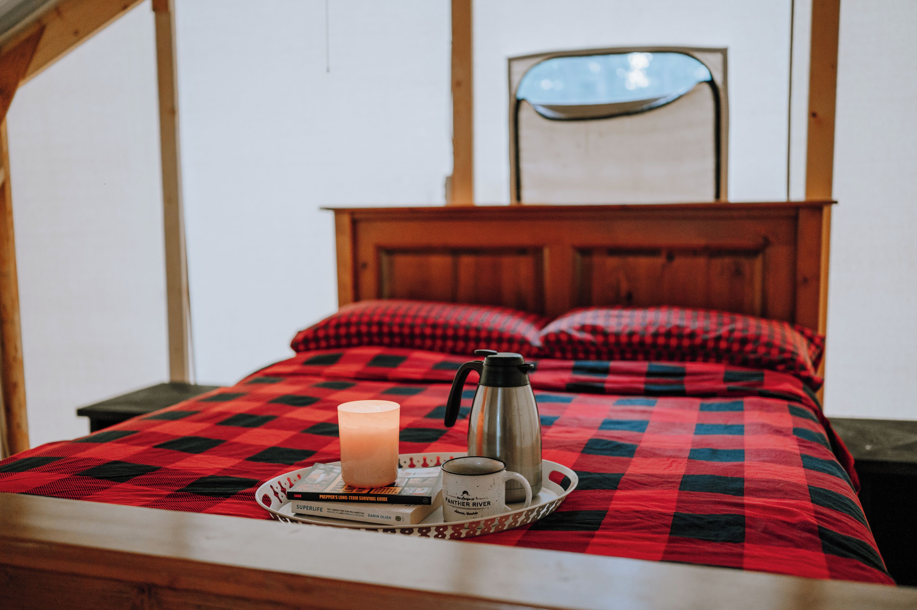 Een bed met een geruit beddegoed, een nachtkastje met een kaars, een koffiezetapparaat en een dienblad met boeken en een mok.