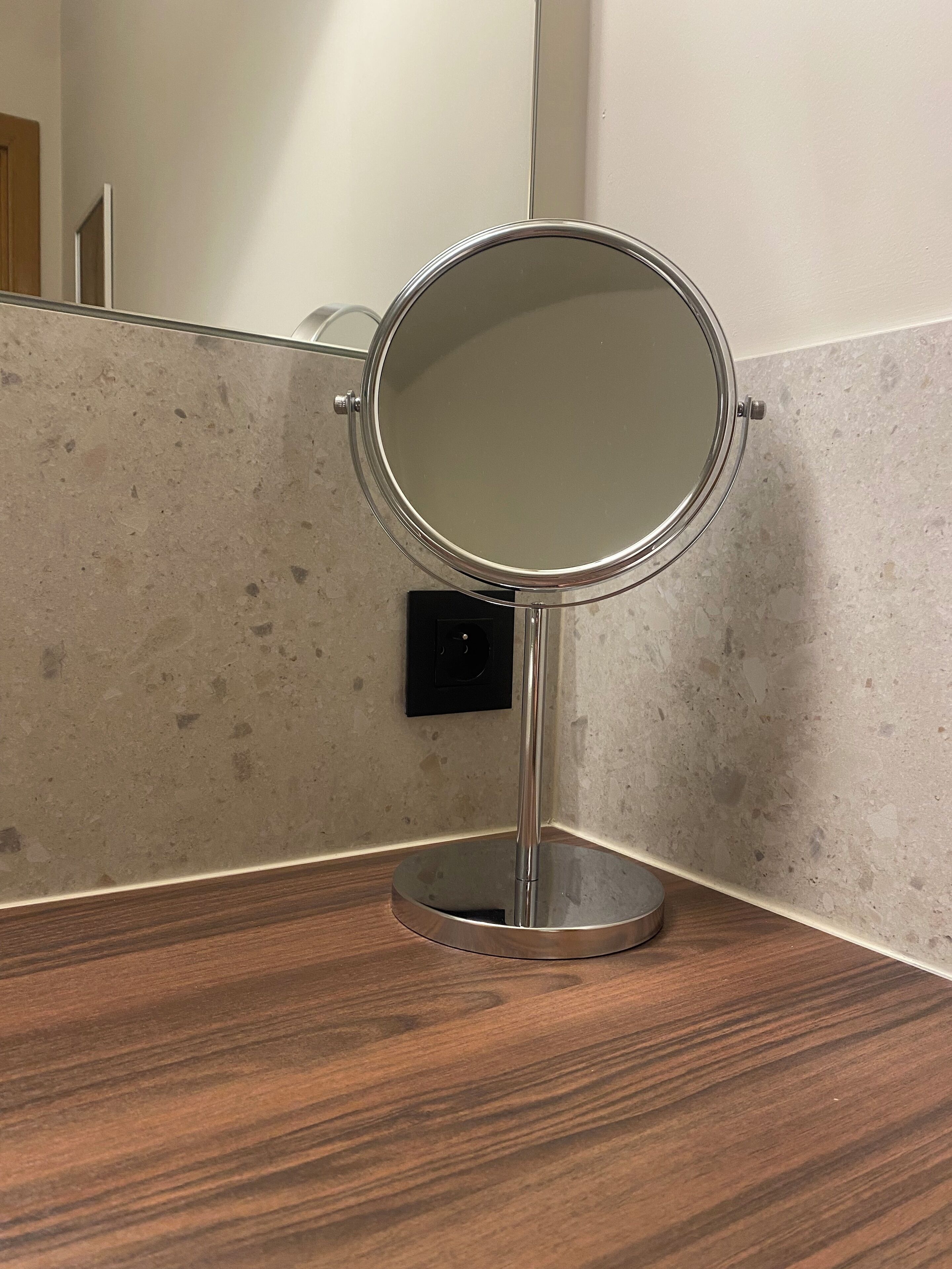 A round mirror on a wooden countertop with a metal stand, against a wall with a textured surface and a door in the background.