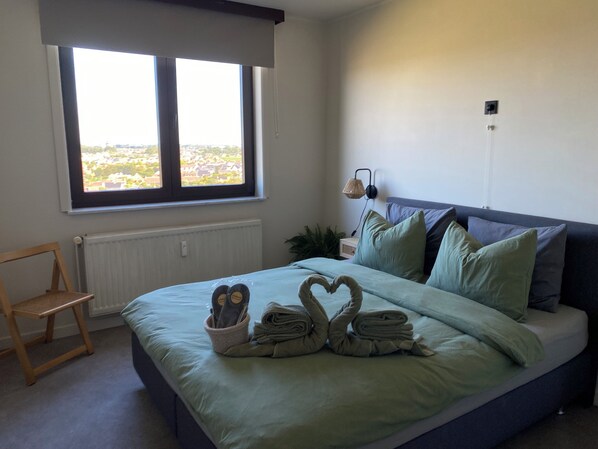 A bedroom with a bed, a chair, a window, and a wall-mounted lamp.