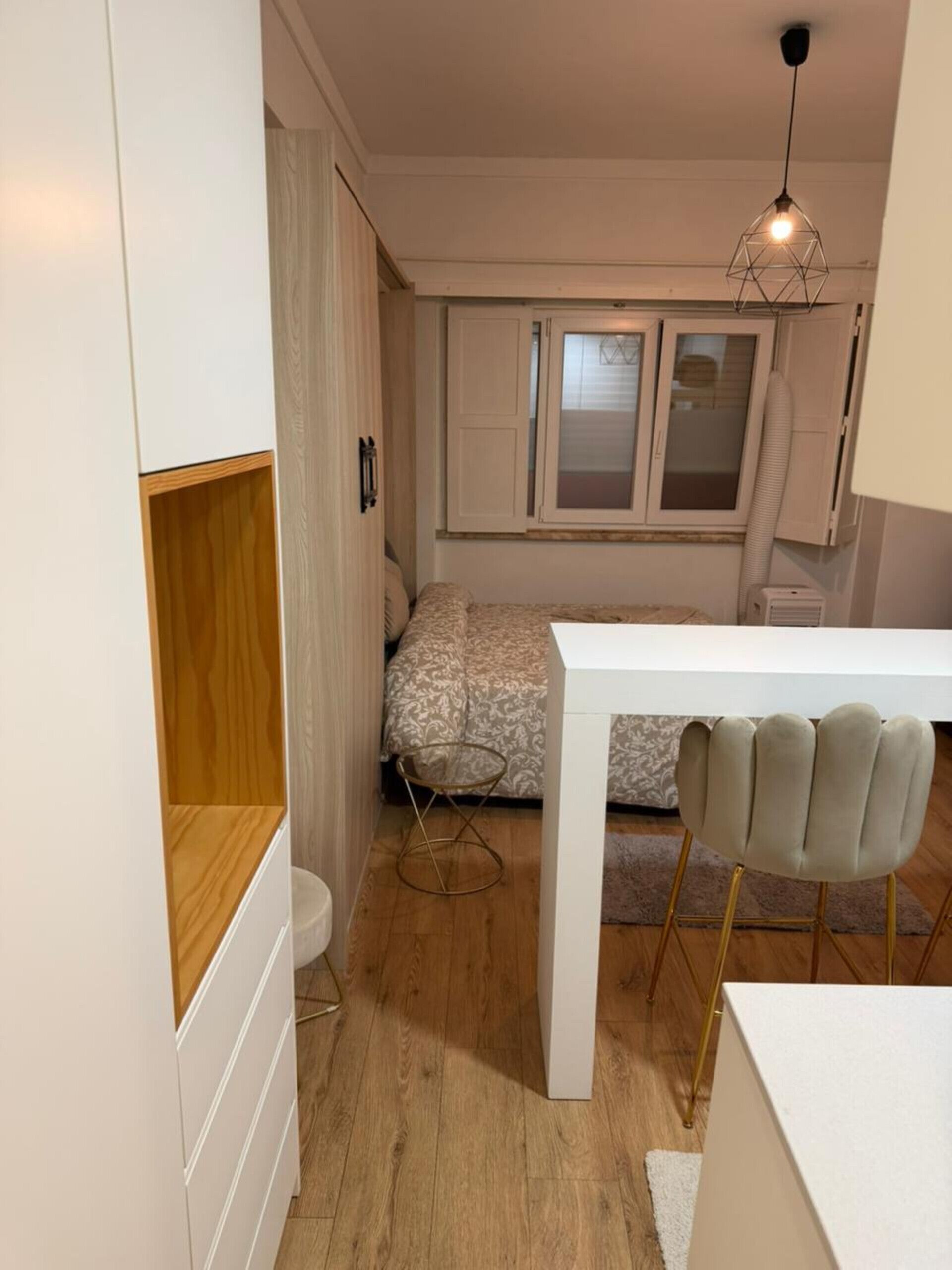 Une pièce compacte et bien éclairée, comprenant un lit, une petite table et une chaise. On y trouve une fenêtre avec des stores, un luminaire au plafond et un radiateur.