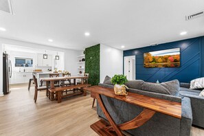 Una cocina moderna con armarios blancos, un comedor con mesa y bancos de madera, un sofá gris y un televisor montado en la pared.