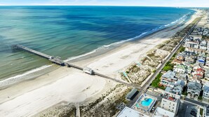 Vue aérienne d’une ville côtière avec une longue jetée qui s’avance dans l’océan, une plage de sable et une rangée de bâtiments le long de la côte.
