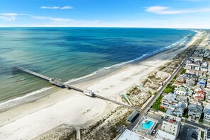 Vista aérea de un pueblo costero con un muelle largo que se adentra en el océano, una playa de arena y una hilera de edificios a lo largo de la costa.