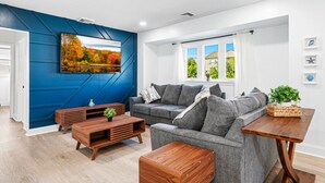Un salon moderne avec un mur d’accent bleu, un canapé gris, une table basse en bois et une fenêtre agrémentée de rideaux blancs.