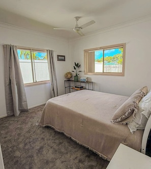 A bedroom with a bed, a ceiling fan, a window with a view, a small table with a globe and books, and a carpeted floor.
