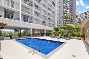 A rooftop swimming pool with a view of high-rise buildings, surrounded by greenery and outdoor seating.
