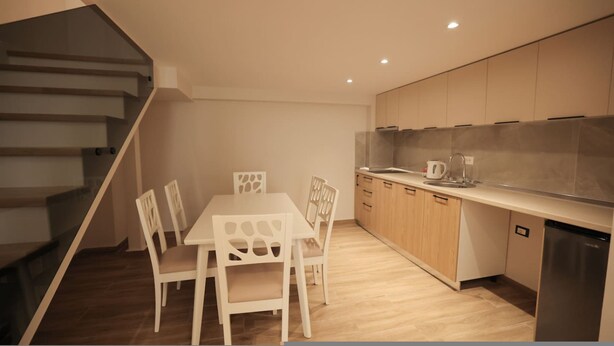 A modern kitchen with wooden cabinets, a stainless steel sink, and a dining area with a white table and chairs. - Duplex City 05 in Durr s (Durrës)