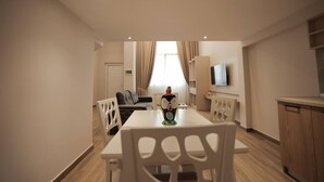 A modern living room with a dining table, white chairs, a sofa, a TV mounted on the wall, and a kitchen area with wooden cabinets. - Duplex City 03 in Durr s (Durrës)