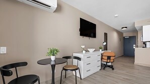 Une chambre d’hôtel moderne équipée d’un téléviseur à écran plat, d’une commode blanche, d’une table de salle à manger ronde avec deux chaises et d’une petite plante en pot.