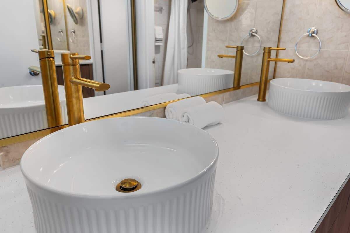 A modern bathroom with a large white sink, gold faucets, and a mirror with a gold frame.