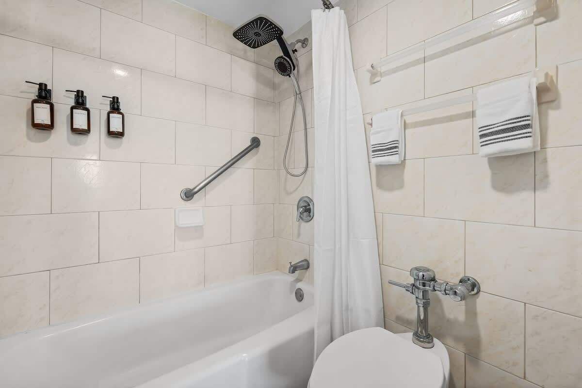 A bathroom with a bathtub, a showerhead, three soap dispensers, a toilet, and a towel rack with towels.