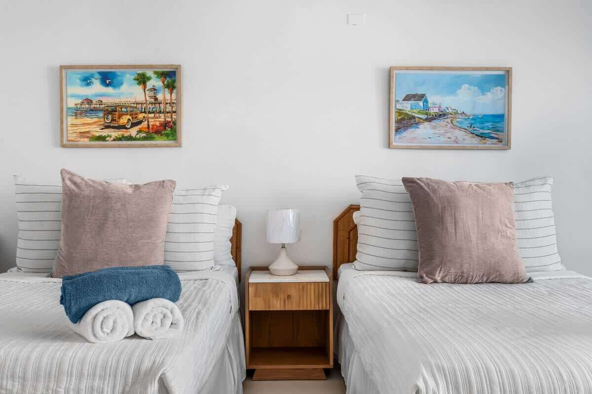 Une chambre d’hôtel avec deux lits, chacun muni d’un oreiller et d’une serviette roulée. Une table de chevet avec une lampe se trouve entre les deux lits. Deux tableaux sont accrochés au mur, au-dessus des lits.
