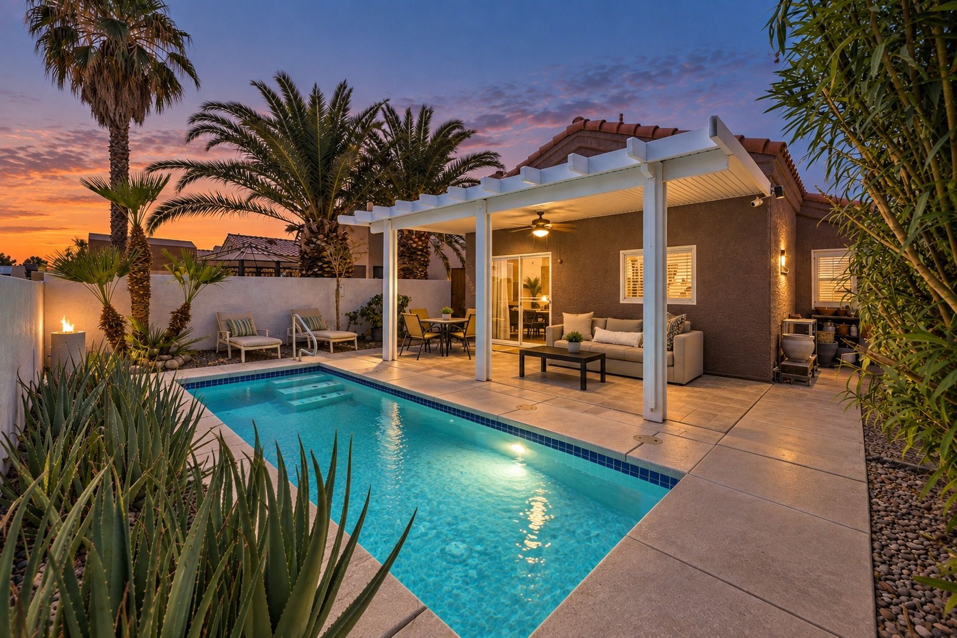 A pool area with a covered patio, seating, and a view of the sunset.