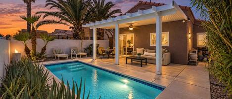 Zona de piscina con un patio cubierto, asientos y vistas al atardecer.