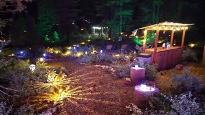 Un espacio al aire libre nocturno con un pabellón de madera, una fogata e iluminación decorativa.