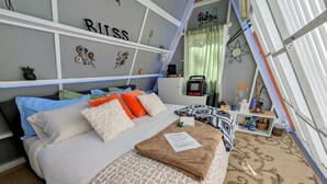 Una cama bien hecha con una colcha estampada, una mesita de noche con un radio, y una pared con detalles decorativos y un cartel que dice 'Welcome'.