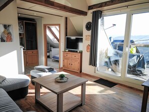 Ein Wohnzimmer mit Fernseher, einem kleinen Tisch mit Pflanze, einer Couch und Blick durch eine Glastür.