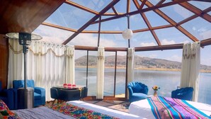 Quarto com uma cama grande, duas poltronas azuis, uma mesinha e vista para um lago através de um teto de vidro.