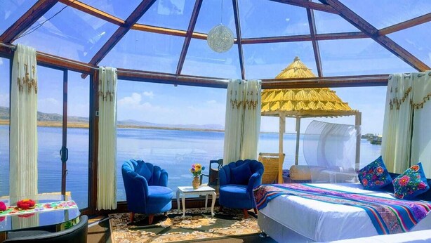 Quarto com uma cama, duas poltronas azuis, uma mesinha com um vaso e vista para a água.