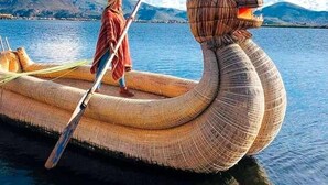 Uma embarcação tradicional com cabeça esculpida, uma pessoa em pé sobre ela e paisagem montanhosa ao fundo.