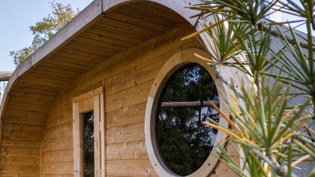 A wooden pod-style cabin with a circular window, a small table with red cushions, and a potted plant. - Wohnung in Niedergurig mit Grill, Sauna und Gart by Interhome (Bautzen)