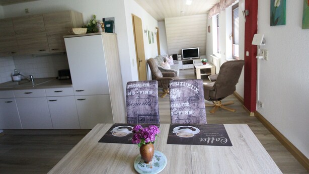 Een moderne keuken met witte kasten, een eethoek met een tafel gedekt voor twee personen, en een woonkamer met een bank en een televisie.