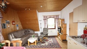 Ein gemütliches Wohnzimmer mit Holzbalkendecke, einer Couch, einem Couchtisch und einem Sessel. Es gibt eine Küchenzeile mit Holzschränken und einer roten Kaffeemaschine.