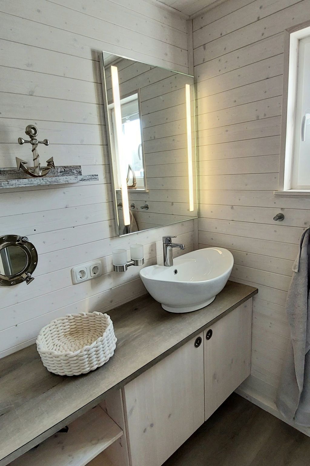 Une salle de bain avec un lavabo sur colonne blanc, un miroir rond, une petite étagère avec des objets décoratifs et un panier tressé posé sur le comptoir.