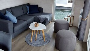 A modern living room with a grey sofa, a white coffee table, and a grey ottoman. There is a painting on the wall and a window with a view of the sea. - Hausboot Tosca by Interhome (Stralsund)
